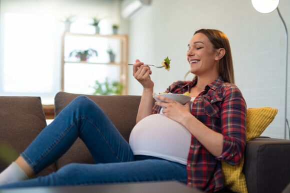 bella-giovane-donna-incinta-felice-che-si-siede-e-che-mangia-macedonia-sul-sofa-a-casa