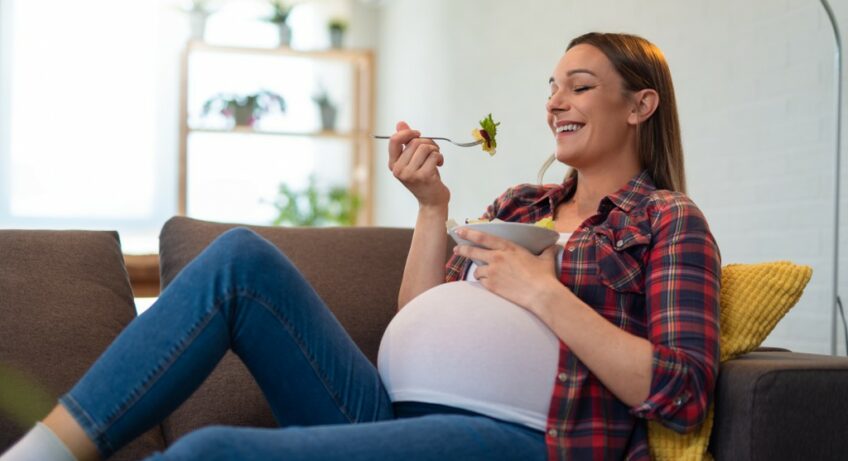 bella-giovane-donna-incinta-felice-che-si-siede-e-che-mangia-macedonia-sul-sofa-a-casa